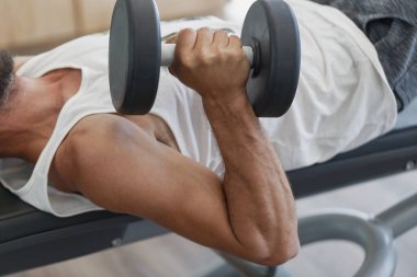 Dambıllarla ağırlık kaldıran erkek sporcu güçlü pompalanmış pazılarını gösteriyor. Kas sporcusu bir bankta uzanıyor, bir spor salonunda göğsüne yakın bir dambıl tutuyor ve bisepsler için egzersiz yapıyor. Adam ağırlık kaldırmak için çaba sarf ediyor.