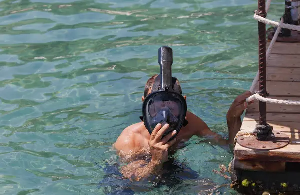 A caucasian man in water next to a wooden dock, wearing with the  black snorkel full face mask diving above the water in sea. One of their hands is holding onto the side of the dock. Swimming goggles
