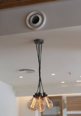 Çoklu ampullü modern avize. Ampuller yanar ve tavana siyah kablolar asılır. Avizenin üzerinde dairesel bir havalandırma deliği görülebilir. Tavan beyaz. Edison 'un ampulü elektrik akımından yanıyor.