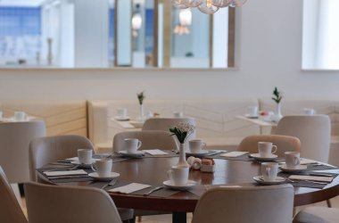 Zarif bir yemek alanı, narin bir avizeyle süslenmiş doğal ışıkla doludur. Yuvarlak masa, beyaz bir çiçek gösteren küçük bir vazoyla tamamlanmış, örülmüş tabaklar ve beyaz fincanlarla dolu. Bej kaplama sandalyeler