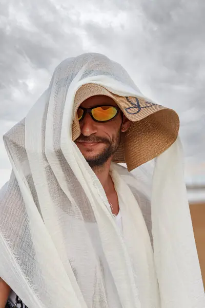 A man wearing sunglasses and a woven womens hat topped with a white scarf. He gazes at the camera, his smile bright and inviting. Behind him, a cloudy sky looms over the sandy earth. Smiling funny men