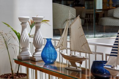 Güzel dekore edilmiş cam masa ve detaylı bir yelkenli. İki bükülmüş mum tutacağı, canlı mavi vazolar ve yeşil bitkiler. Arka plan lüks bir iç mekan rahat bir oturma alanı