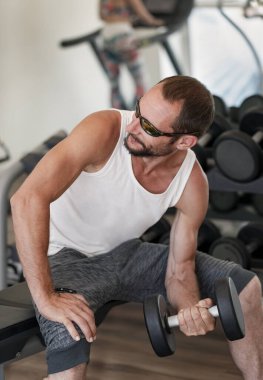 Spor salonunda beyaz atlet, güneş gözlüğü ve koyu renk şort giyen bir dambılı kaldıran beyaz bir adam. Sakallı, spor salonunda ağırlık kaldıran, yana bakan güçlü bir adam. Ağırlık tutan adama yaklaş.