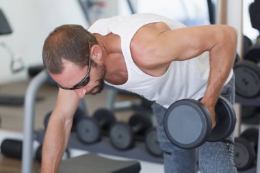 Parlak ışıklı bir spor salonunda kaslı, beyaz, güçlü bir adam dambıl kürek çekiyor. Kas çalışan odaklanmış bir adam, tek kollu halterli sıra yapıyor. Beyaz bir atlet ve güneş gözlüğü takıyor..