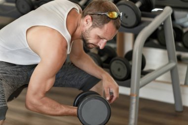 Kafkasyalı bir adam fitness kulübünün rafına yakın bir yerde oturuyor ve dambılı kaldırıyor. Spor salonunda biceps kıvıran erkek ağırlık antrenörü. Olgun bir adam spor salonunda çalışırken dambıl kullanıyor.