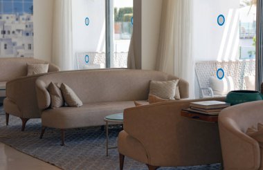 Bej koltukları, cam sehpası ve dokuma halısı olan modern bir oturma odası. Oda peluş yastıklar ve kitaplarla süslenmiş. Geniş pencereler, alanı ışıkla aydınlatan bir dış görünüm ortaya çıkarır.