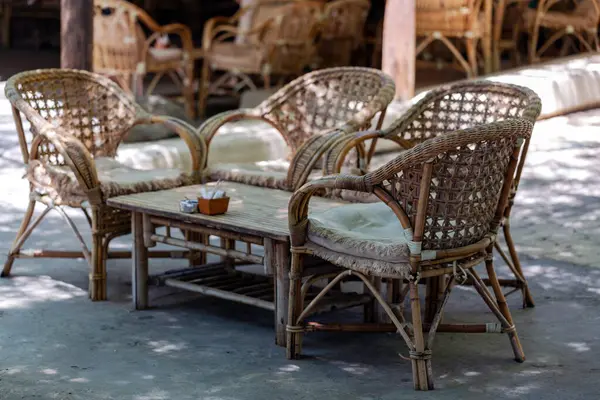 Outdoor setting with three wicker chairs and a matching table adorned with cushions. The furniture is arranged on a concrete floor under the shade, creating an inviting atmosphere for relaxation