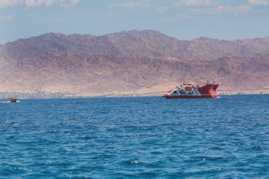 Kırmızı ve beyaz bir tekne sakin mavi sularda yüzer. Arka planda, engebeli kahverengi dağlar dingin denizle çok güzel tezat oluşturuyor. Gökyüzü açık, sahnenin genel sakinliğine huzur katıyor..