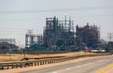  Tüten bacaları olan Ölü Deniz tuzları üretimi için bitki. Metal kirişler, borular ve kulelerden oluşan karmaşık bir ağ görünür. Temiz bir gökyüzüne karşı dumanlar yükseliyor. Yol uzanıyor