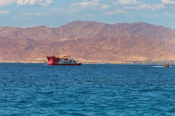 Stock image Red and white modern boat sailing on a tranquil blue sea. The backdrop features rugged mountains under a clear sky. The mountains, though barren, add a majestic element to the scene. Red sea, Israel