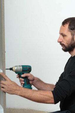 Craftsman in dark clothing drilling a power drill and screw collects wooden furniture at home. Close up of man with green and black electric hand drill, power tool, screwdriver against a white wall clipart