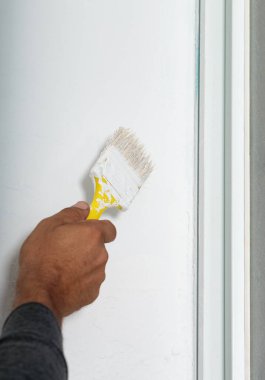 Duvarı beyaza boyayan ve sarı saplı boya fırçası kullanan bir adam. Fırçanın kılları beyaz boyayla kaplıdır. Duvar, görünür çerçevenin gösterdiği gibi bir pencerenin yanında.