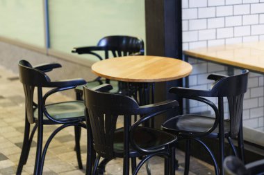 Yuvarlak ahşap bir masanın etrafında dört siyah sandalye. Sandalyelerin arkası açık modern bir tasarımı var. Beyaz fayanslar arka plandaki duvarı kaplıyor. Zemin kare karolardan yapılmıştır. Teras açık havada.