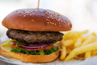 Ekmeğinde susam tohumu olan bir hamburger yakın çekim. Yanında altın patates kızartması var. Hamburgerde marul, domates, turşu ve kırmızı soğan var. İştah açıcı ve görsel olarak çekici görünüyor..
