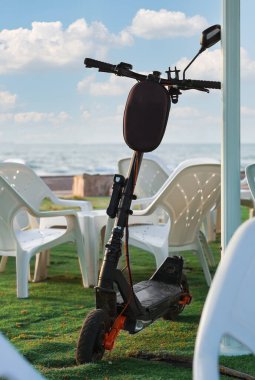 Güneşli bir günde deniz kenarında park etmiş elektrikli scooter. Siyah elektrikli scooter beyaz bir masanın yanına park edilmiş ve arka planda mavi gökyüzü ve sakin deniz manzaralı sandalyeler var.. 