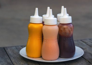 Lezzetli sos şişeleri sarı, turuncu ve kırmızı renkli beyaz bir tabakta ahşap masa zemininde bulunur. Hardal, mayonez ve ketçap içeren şişeler, beyaz kapaklı soslu barbekü.