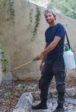 Haşere kontrolü. Koyu mavi tişörtlü ve siyah pantolonlu bir adam sırtında uzun bir hortum bulunan sprey bir alet kullanıyor. Adam yeşil yapraklarla taş basamaklarda duruyor.
