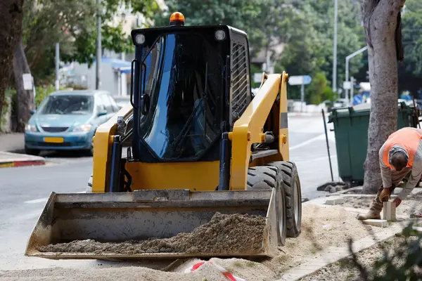Kovası indirilmiş ve pislikle dolu bir caddede sarı buldozer. Turuncu yelekli bir işçi eğilmiş kaldırımda çalışıyor. Arka planda ağaçlar, park edilmiş arabalar var. İnşaat