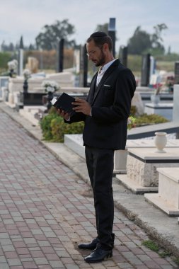 Sakallı ve kippah giyen bir Yahudi Tevrat 'tan ölenler için dua okuyor. Resmi takım elbiseli Yahudi erkek mezarlıkta duruyor. Mezar taşları ve çiçek aranjmanları arasında.
