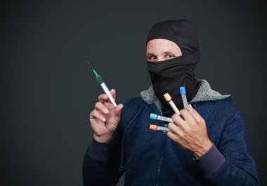 Mavi kapüşonlu ve siyah maskeli bir adam bir elinde şırınga, diğer elinde de birkaç kan toplama tüpü tutuyor. Sadece gözleri görünür, yoğun ve odaklanmış bir bakış verir. Karanlık arkaplan