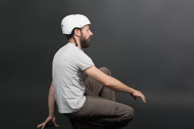 A bearded man wearing a white helmet sits in profile against a dark background. He is dressed casually in a white t-shirt sits in a balanced pose showcasing his protective gear against grey background clipart