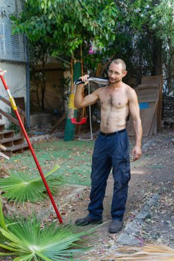Dışarıda sakallı, tişörtünü omzunun üstünde tutan tişörtsüz bir adam. Yanında kırmızı bahçe aletleri ve yere saçılmış palmiye yaprakları olan koyu renk pantolon ve botlar giyiyor. Açık hava işi.