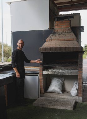 Man stands by a large outdoor brick barbecue grill, dressed in dark clothing gestures towards the grill. Bags of charcoal are stored underneath. Empty burning grill in a backyard with artificial grass clipart