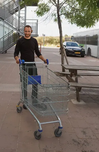Günlük kıyafet giymiş gülümseyen bir adam boş bir alışveriş arabasını bir bankın ve otoparkın yakınına itiyor. Ayar, arka planda park halindeki arabaların bulunduğu ticari veya kentsel bir alanı yansıtıyor.
