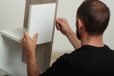 Bir adam bir mobilya parçasını birleştirmeye odaklanır, panelleri hassasiyetle bir araya getirir. O, iyi ışıklandırılmış bir odada, göreve dikkatli bir yaklaşım gösteriyor. Cihazlar ve malzemeler yakında.