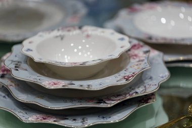 Bir yığın zarif porselen tabak ve bir kase, her birinde pastel renklerle karışık çiçek desenleri ve altın budanmış kenarlar var. Tabaklar, tabaklar düzgünce katlanmış, istiridyeli jantlar, parlak yüzeyler.