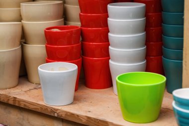 Various ceramic plant pots in different colors are neatly stacked on a wooden shelf at a market. Vibrant colors include red, white, beige, and green, showcasing diverse styles. Colorful ceramic pot clipart