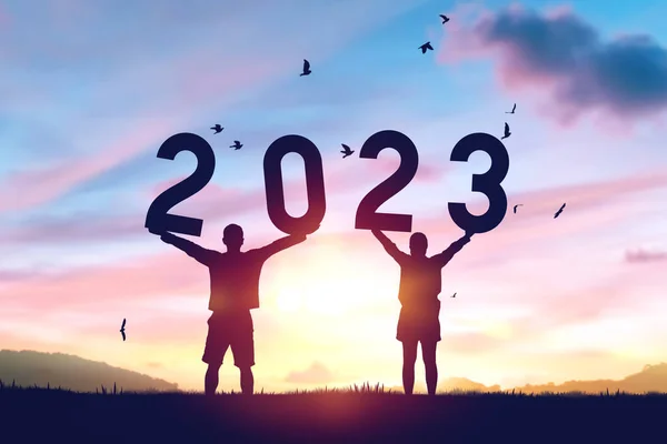 Couple raise hand up and holding number 2023 on sunset sky with birds flying at top of mountain and  abstract background. Happy new year and holiday concept. Vintage tone filter effect color style.