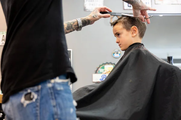 Barber cutting a boys hair with scissors at the barbershop. Hairdressing and childhood concept.
