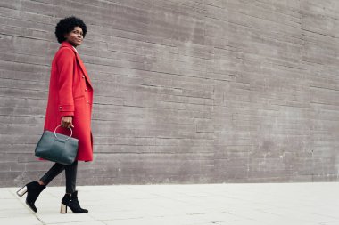 Havalı bir afro kadın elinde çanta ve kırmızı bir palto ile sokakta yürüyor..