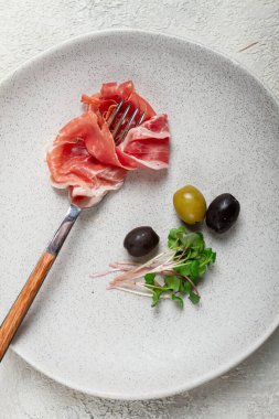 Bir çatalda prosciutto, üst görünüm, zeytin ve tabakta mikro yeşiller, açık arkaplan, insan yok.,