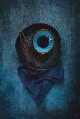 Yemek arkaplanı, boş bir siyah tabak, bir kase, ahşap yemek çubukları, bir peçete, mavi arka planda, masa düzeneği, üst manzara, insan yok.,