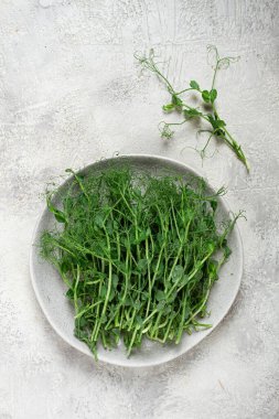 Taze toplanmış bezelye filizi koleksiyonu açık seramik bir tabakta oturur ve ince yeşil yapraklarını ve filizlerini dokulu bir yüzeye karşı sergilerler. Çekimlerden biri yakınlarda duruyor..