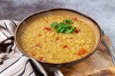 A bowl of warm lentil soup featuring vibrant diced tomatoes and a garnish of fresh herbs rests on a wooden table alongside a striped kitchen towel, ideal for a comforting dining experience. clipart