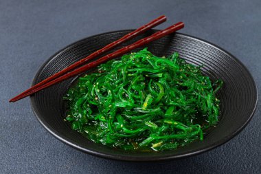 Chuka salatası, taze deniz yosunundan yapılmış, susam soslu, siyah bir kasede servis ediliyor, çubuklarla servis ediliyor.,
