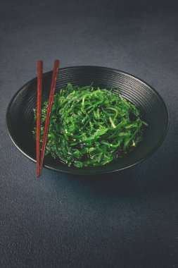Chuka salatası, taze deniz yosunundan yapılmış, susam soslu, siyah bir kasede servis ediliyor, çubuklarla servis ediliyor.,