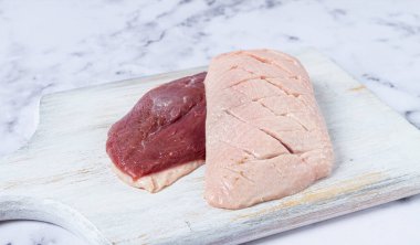 Taze çiğ ördek göğsü, tahta bir kesme tahtasının üzerine serilmiş, beyaz mermer bir yüzey üzerinde.
