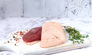 Taze çiğ ördek göğsü, ahşap bir kesim tahtasına yayılmış, beyaz mermer bir yüzey üzerinde otlar ve baharatlarla,