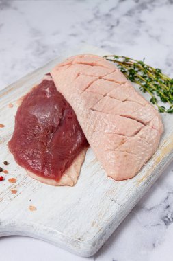 Taze çiğ ördek göğsü, ahşap bir kesim tahtasına yayılmış, beyaz mermer bir yüzey üzerinde otlar ve baharatlarla,