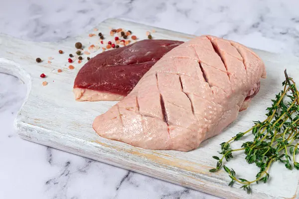 Taze çiğ ördek göğsü, ahşap bir kesim tahtasına yayılmış, beyaz mermer bir yüzey üzerinde otlar ve baharatlarla,