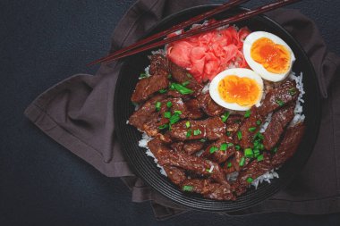 Udon Japon bifteği, pilav, zencefil turşusu, haşlanmış yumurta dilimi, yeşil soğanla süslenmiş, siyah bir kasede, çubuklarla.,