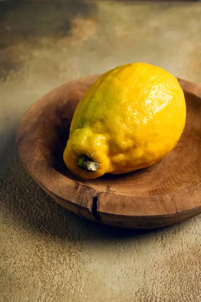 Canlı sarı bir limon elle oyulmuş ahşap bir kasede oturur. Limonun desenli teni doğal ışıkta parlıyor, rengini ve tazeliğini kırsal zemine vurguluyor..