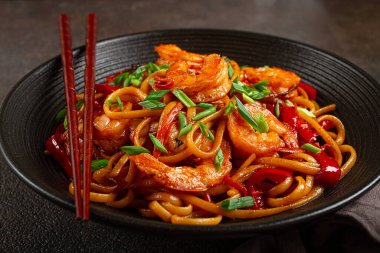 Kırmızı dolmalık biber ve yeşil soğanlı udon kızartmalı karides eriştesi, siyah bir kasede servis ediliyor, çubuklarla servis ediliyor, koyu arka plan...,