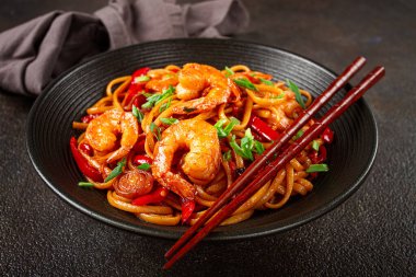 Kırmızı dolmalık biber ve yeşil soğanlı udon kızartmalı karides eriştesi, siyah bir kasede servis ediliyor, çubuklarla servis ediliyor, koyu arka plan...,