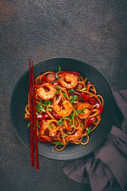 Kırmızı dolmalık biber ve yeşil soğanlı udon kızartmalı karides eriştesi, siyah bir kasede servis ediliyor, çubuklarla servis ediliyor, koyu arka plan...,
