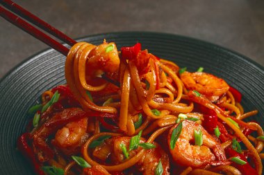 Kırmızı dolmalık biber ve yeşil soğanlı udon kızartmalı karides eriştesi, siyah bir kasede servis ediliyor, çubuklarla servis ediliyor, koyu arka plan...,